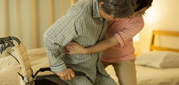 Stock photo of one person lifting another out of a wheelchair with a bed in the background.
