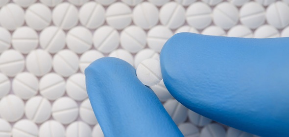 Background of round white pills with an indented line through the center. From the bottom right of the image a thumb and index finger in blue latex gloves extend holding another white pill with an indented line through the middle. 