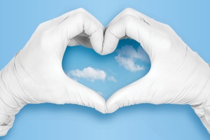 Photo of medical gloved hands making a heart shape against a blue sky.